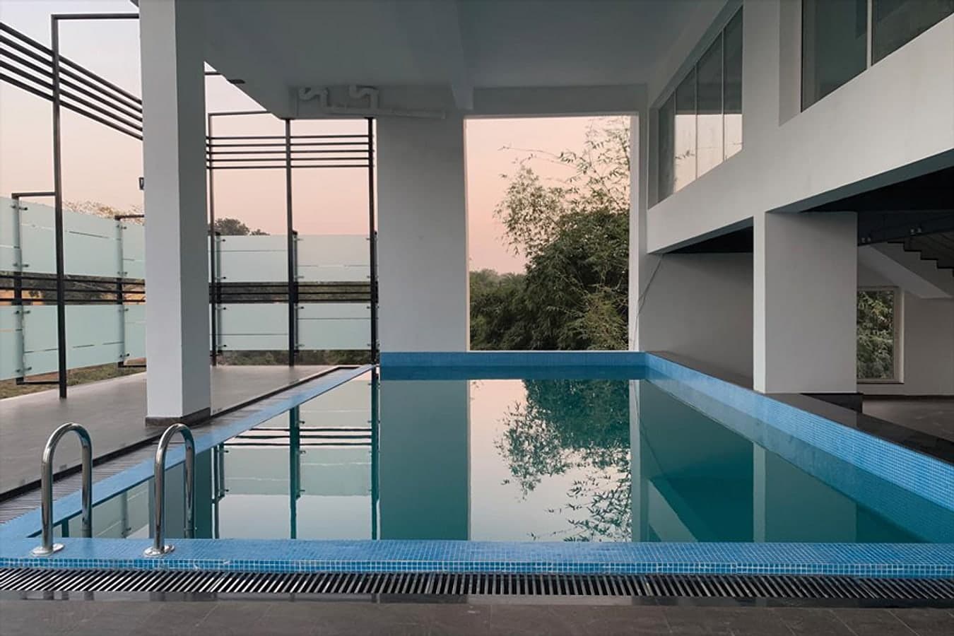 Blue-tiled swimming pool at dusk reflecting trees and a pink sky between white pillars