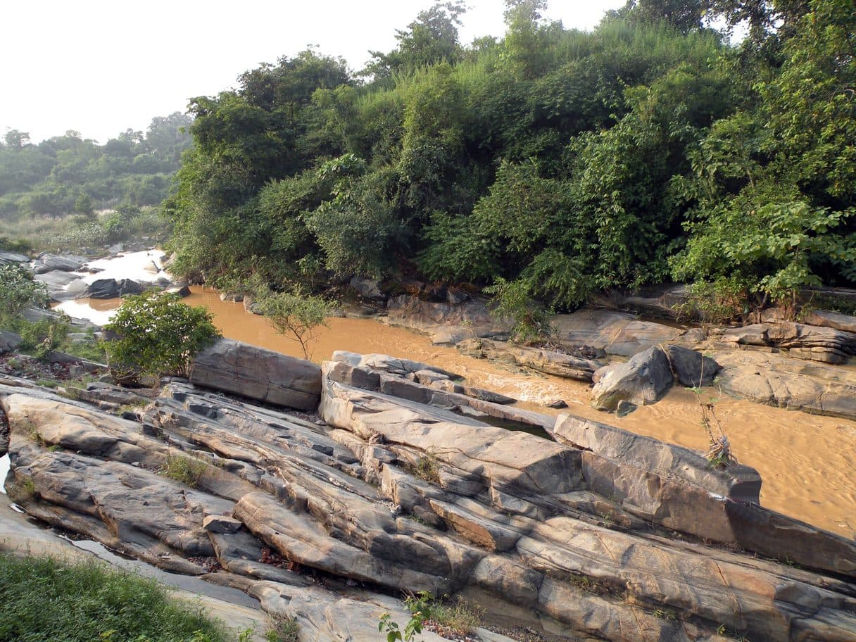 The rocky bed of the Barakar river behind Kalyaneshwari temple, with brown water and dense green trees